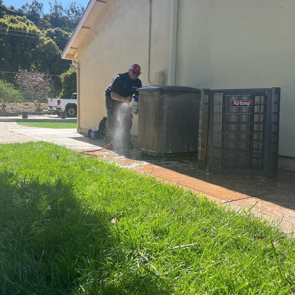 Worker in a cap spraying water beside outdoor HVAC units next to a beige building.