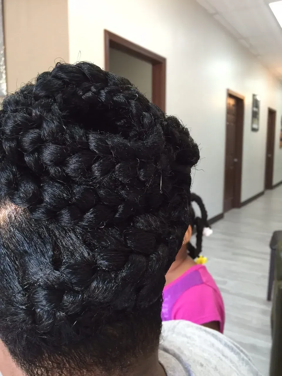Person with an intricate black braided updo in a hallway, with another person in a pink top behind them