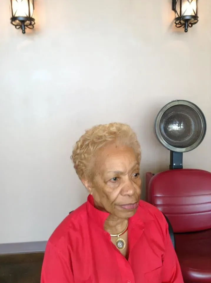 Person in a red shirt seated indoors, looking to the side, with wall lights and a fan in the background.