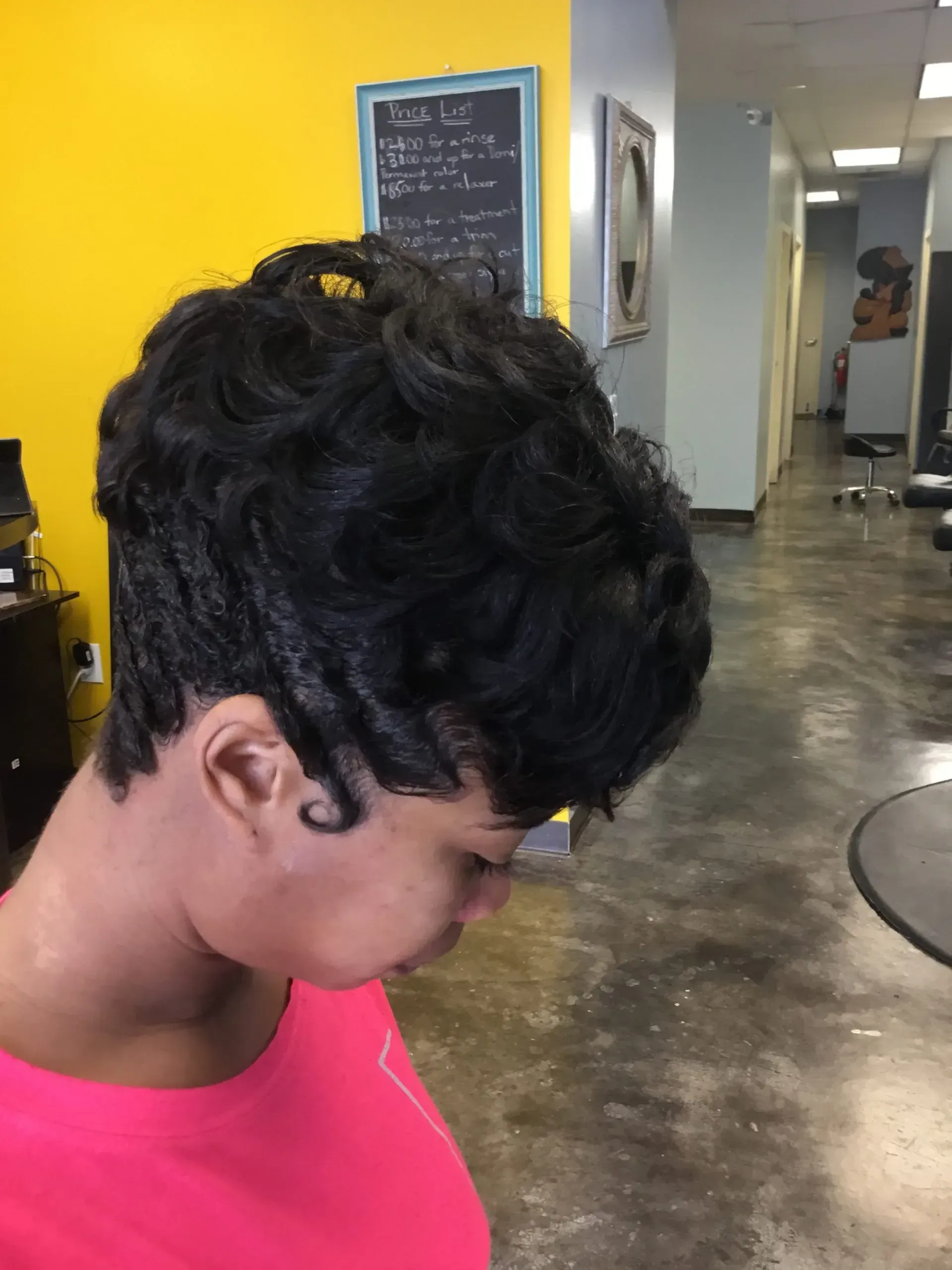 Person in a pink top looking downward in a salon with yellow walls and a mirror.