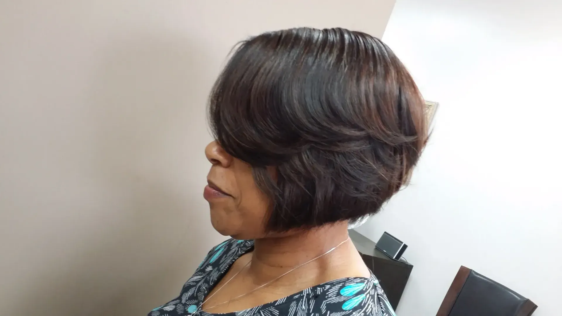 Woman in profile with a short dark bob hairstyle, seated against a light wall.