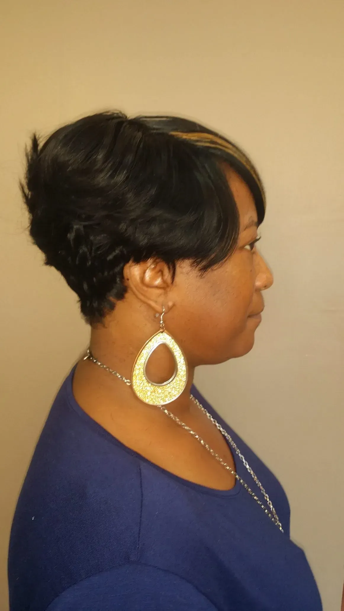Side profile of a woman with short dark hair, wearing a blue top and large gold hoop earrings against a beige background.