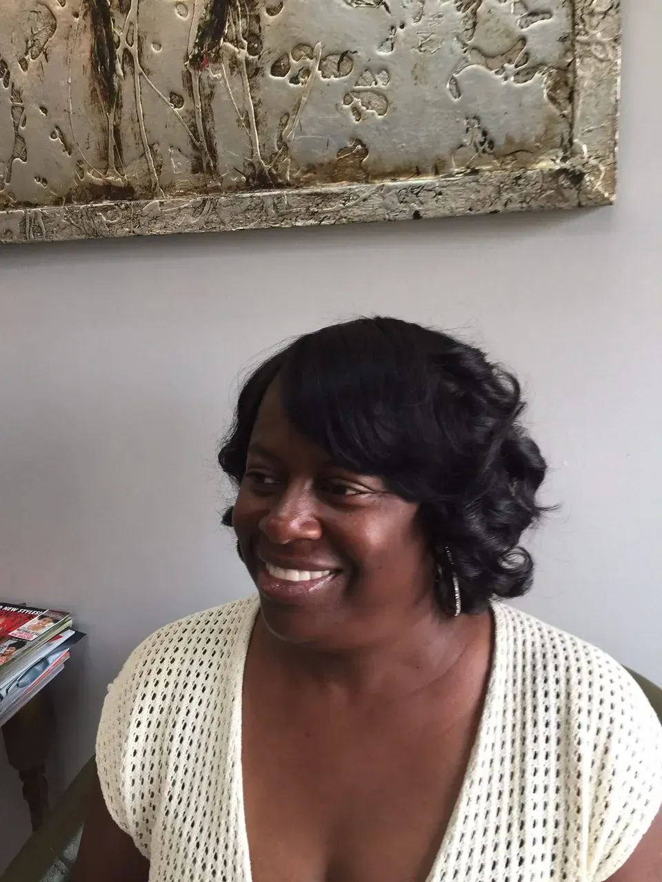 Smiling woman seated indoors beside wall art, wearing a cream knit top