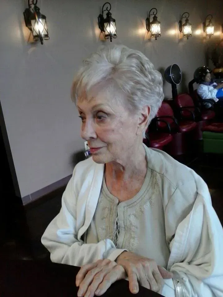 Older woman in a cream sweater seated indoors, looking to the side with wall sconces behind her