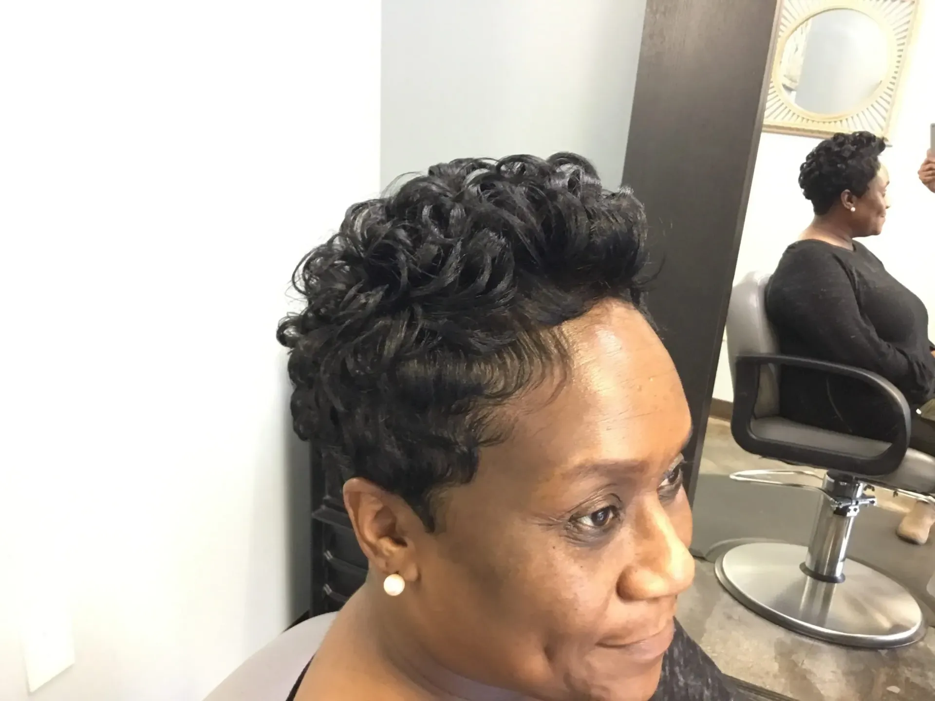 Person with short curly hair seated in a salon chair, shown in side profile with a mirror reflection.