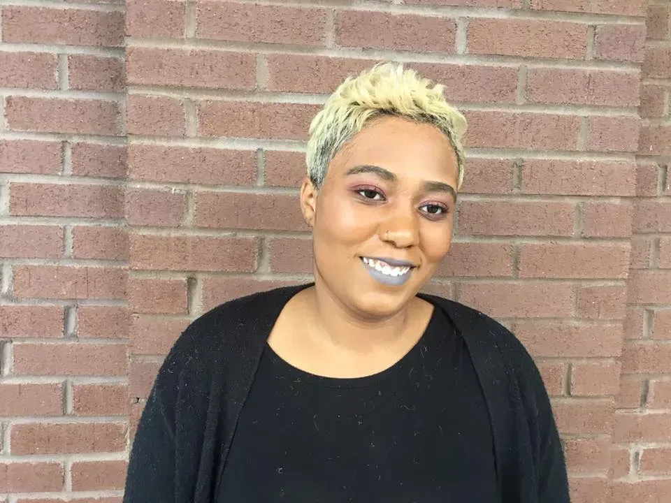 Smiling person with short blond hair and blue lipstick standing in front of a brick wall