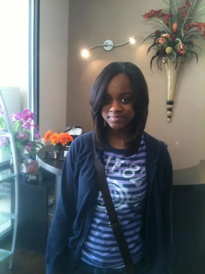 Woman standing indoors by a window, wearing a striped shirt and navy cardigan, with flowers and wall decor behind her