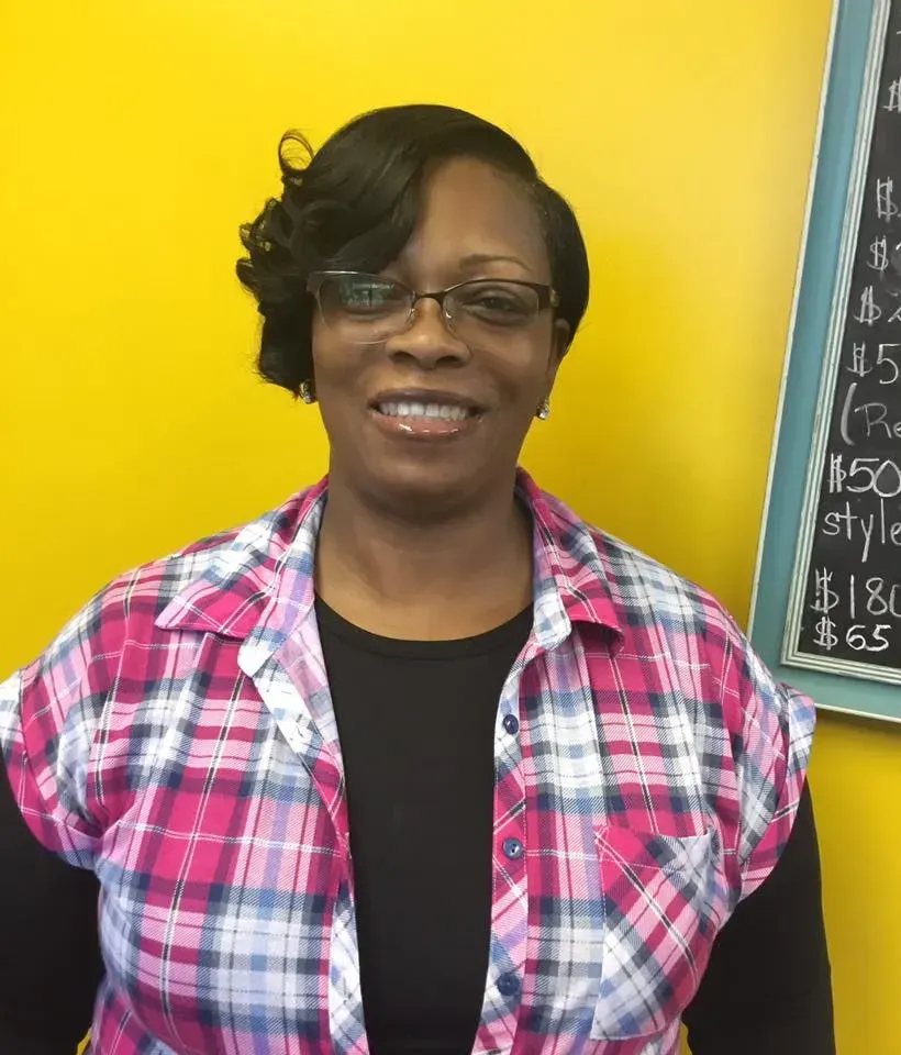 Smiling woman in glasses and plaid shirt standing before a bright yellow wall and chalkboard.