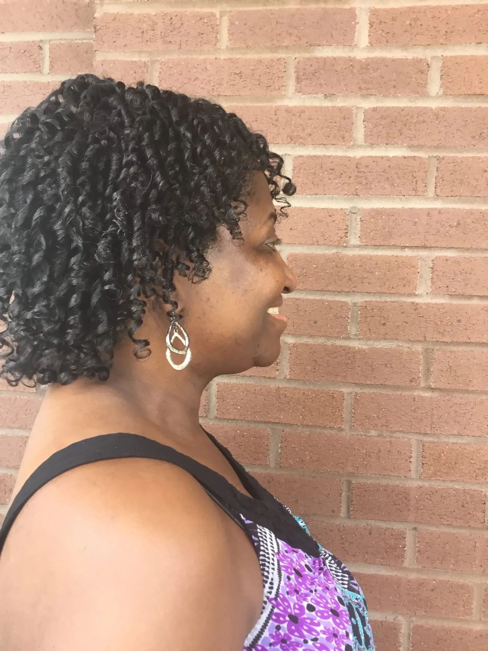 Profile view of a woman with curly hair and hoop earrings, standing against a brick wall in a purple patterned top