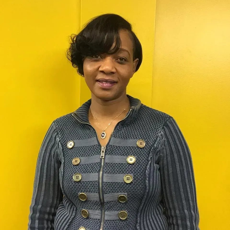 Woman standing against a bright yellow wall, wearing a dark gray jacket and smiling softly.