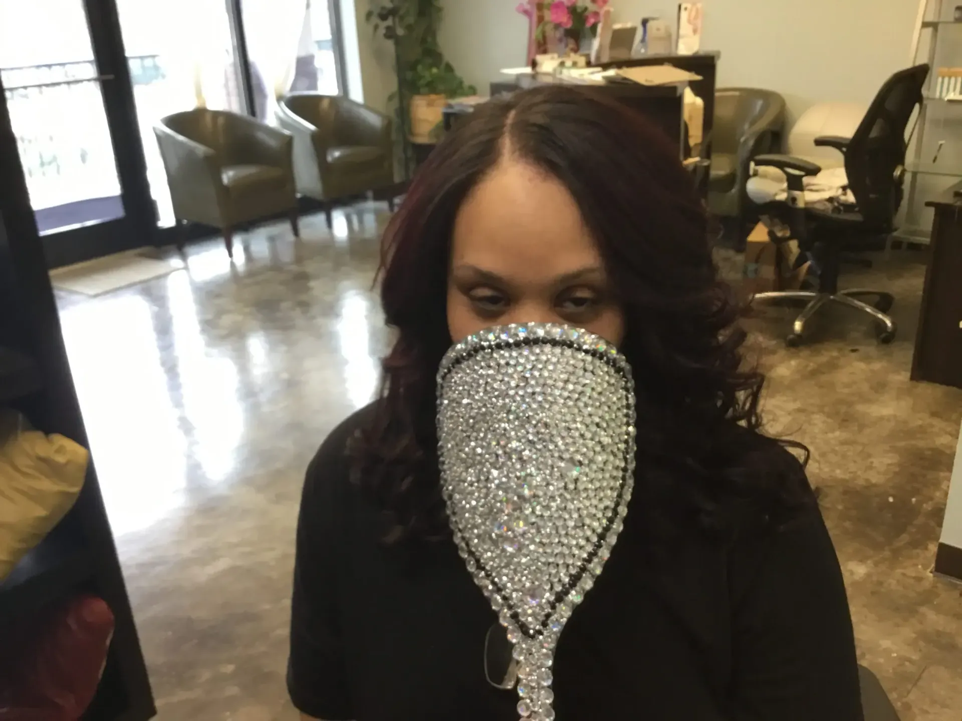 Person in a salon wearing a sparkling face covering, seated in a chair with styling stations in the background