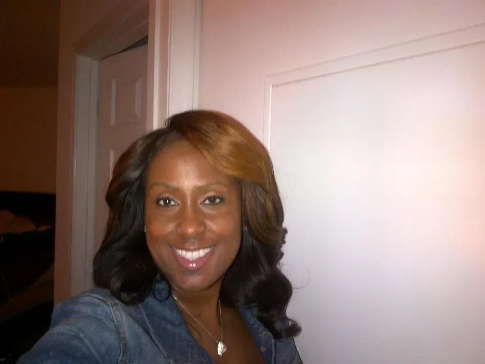 Smiling person with shoulder-length hair in a denim jacket standing indoors near a doorway