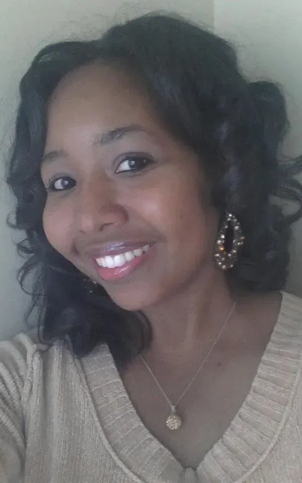 Smiling person in a beige sweater, wearing dangling earrings and a necklace, posing indoors.