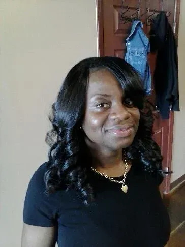 Woman with curled black hair and black shirt smiling indoors near a doorway and hanging clothes