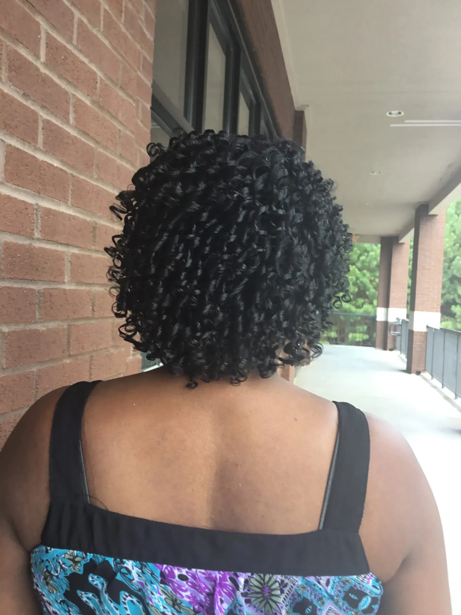 Back view of a person with short curly hair walking beside a brick building and covered walkway
