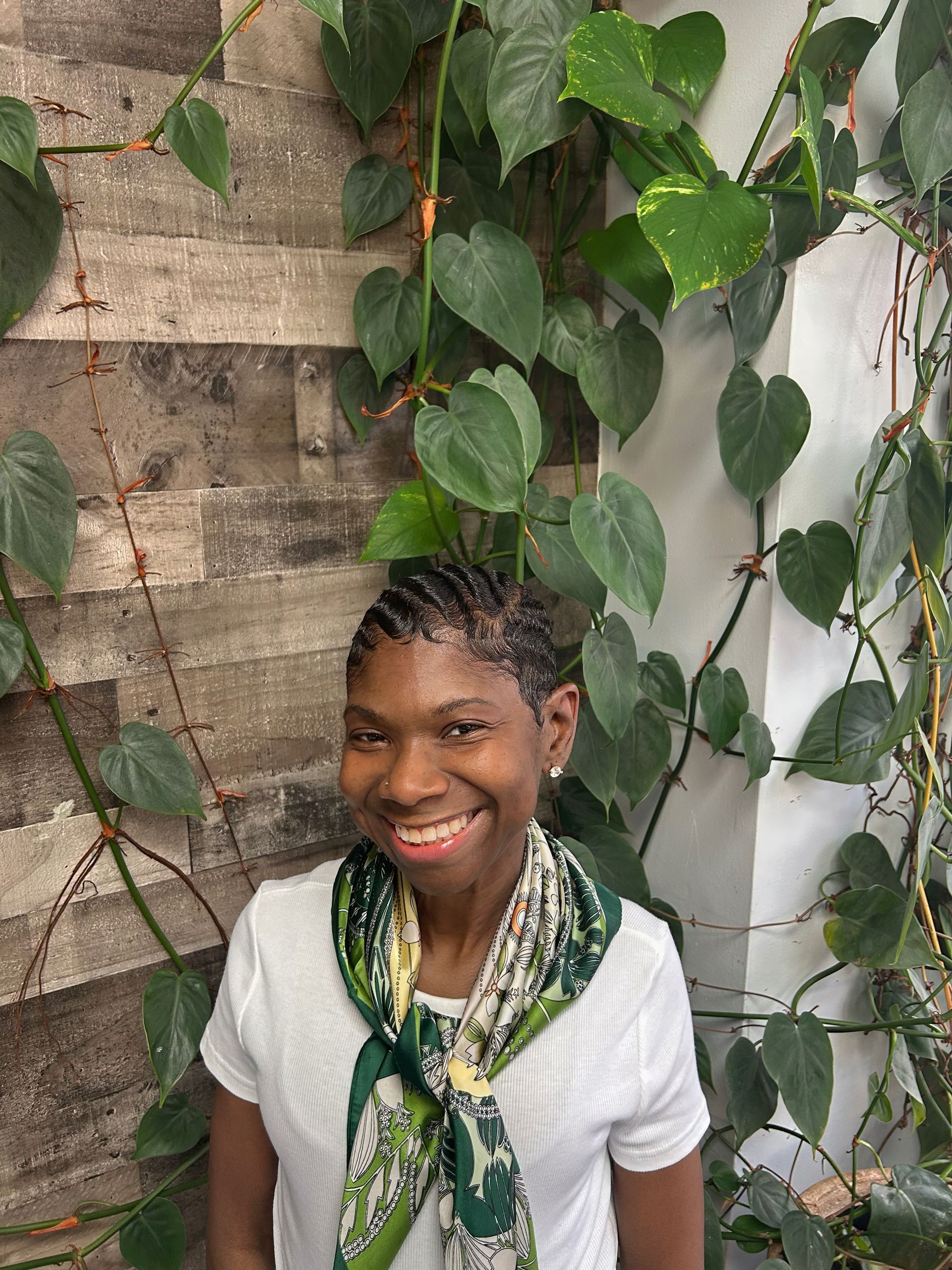 Smiling person in a white shirt and green scarf standing beside a leafy wall with stone tiles