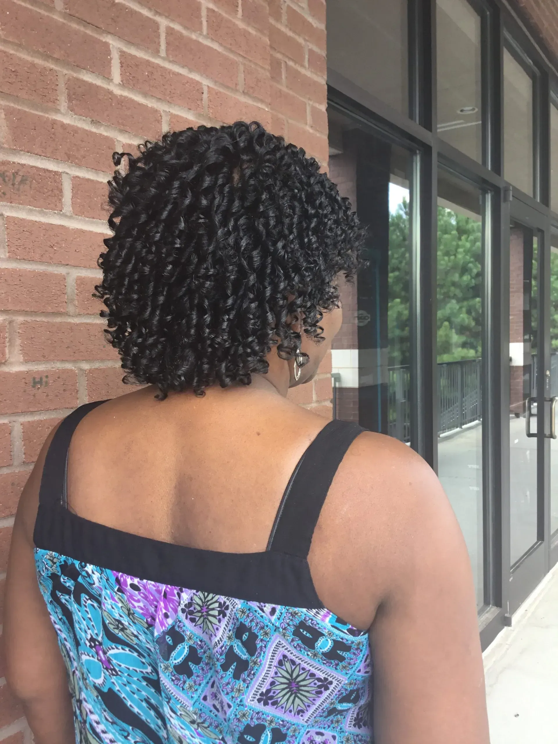 Person with curly hair wearing a colorful top, seen from behind beside a brick wall and windows.