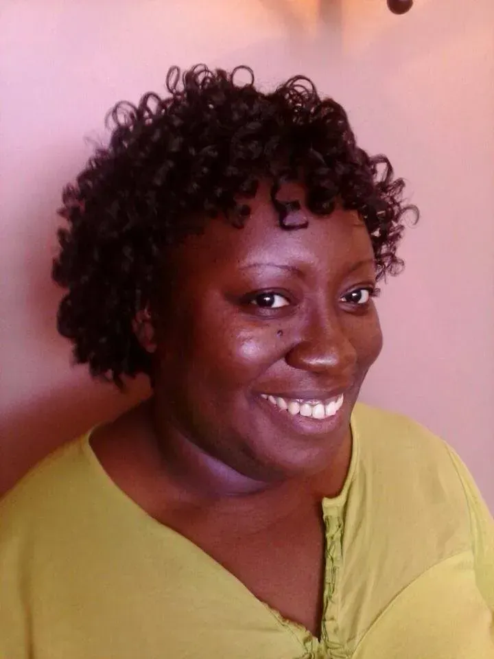 Smiling person with curly hair wearing a yellow top against a pink wall