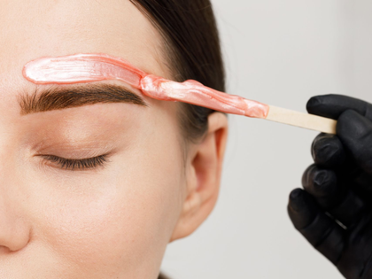 Eyebrow waxing with pink wax applied using a wooden spatula on a closed eye area