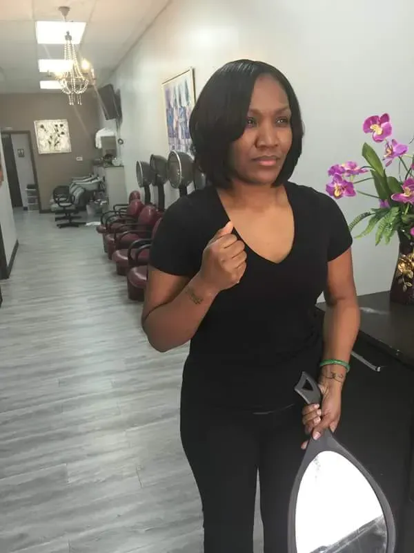 Woman standing in a salon hallway, holding a phone and a bag, with purple flowers in the foreground