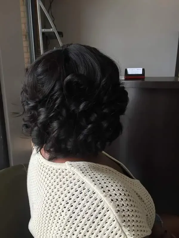 Person with short curly black hair seated indoors, viewed from behind in a light knit top.