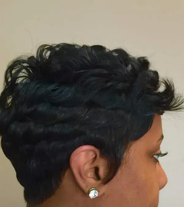 Close-up side profile of a person with short black curly hair and a silver stud earring against a beige wall