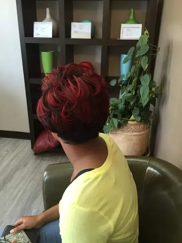 Person with red hair seated in a living room, looking toward shelves and a potted plant.