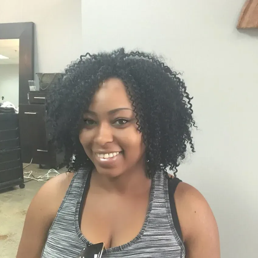 Smiling woman with curly black hair in a gray tank top, standing indoors in a salon.