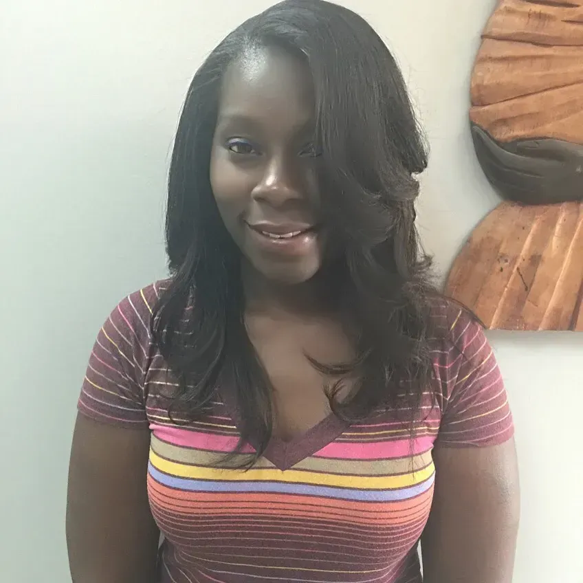 Smiling woman in a striped shirt with long dark hair, standing indoors against a light wall.