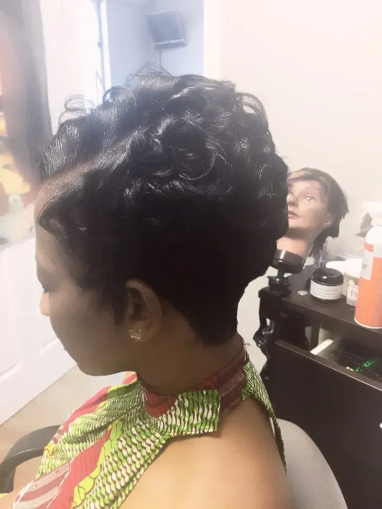 Woman in a green patterned top with short curly hair, seated in a salon chair facing left.