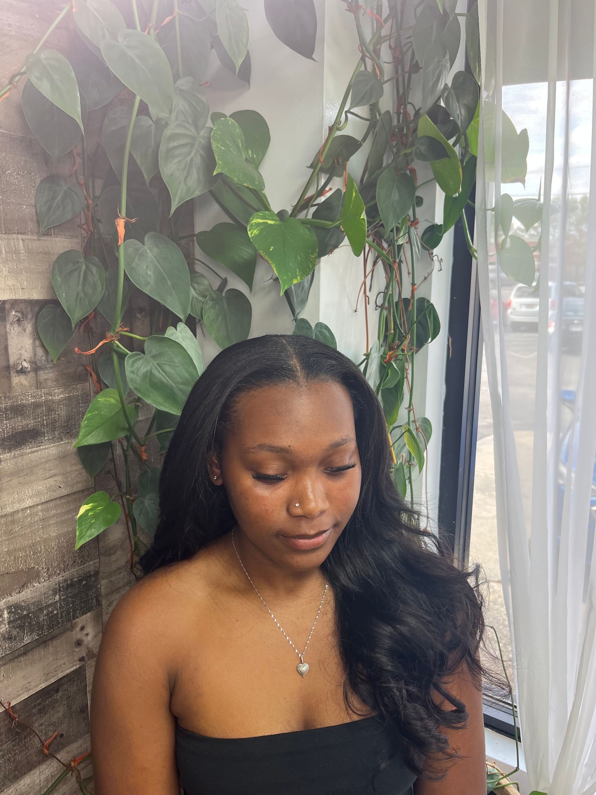 Woman with long black hair sitting beside lush green plants near a window, looking down thoughtfully