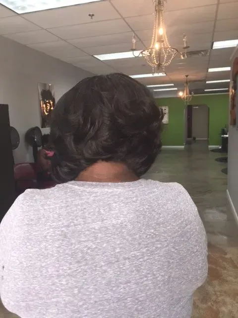 Person seen from behind in a salon, with dark hair and a gray top, facing mirrors and salon chairs.