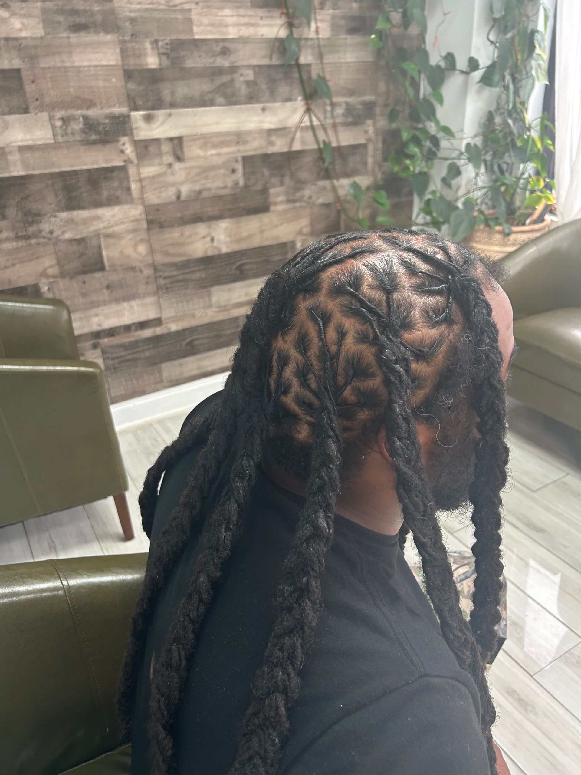 Person with long braided hair seated in a chair beside a window and textured wall.