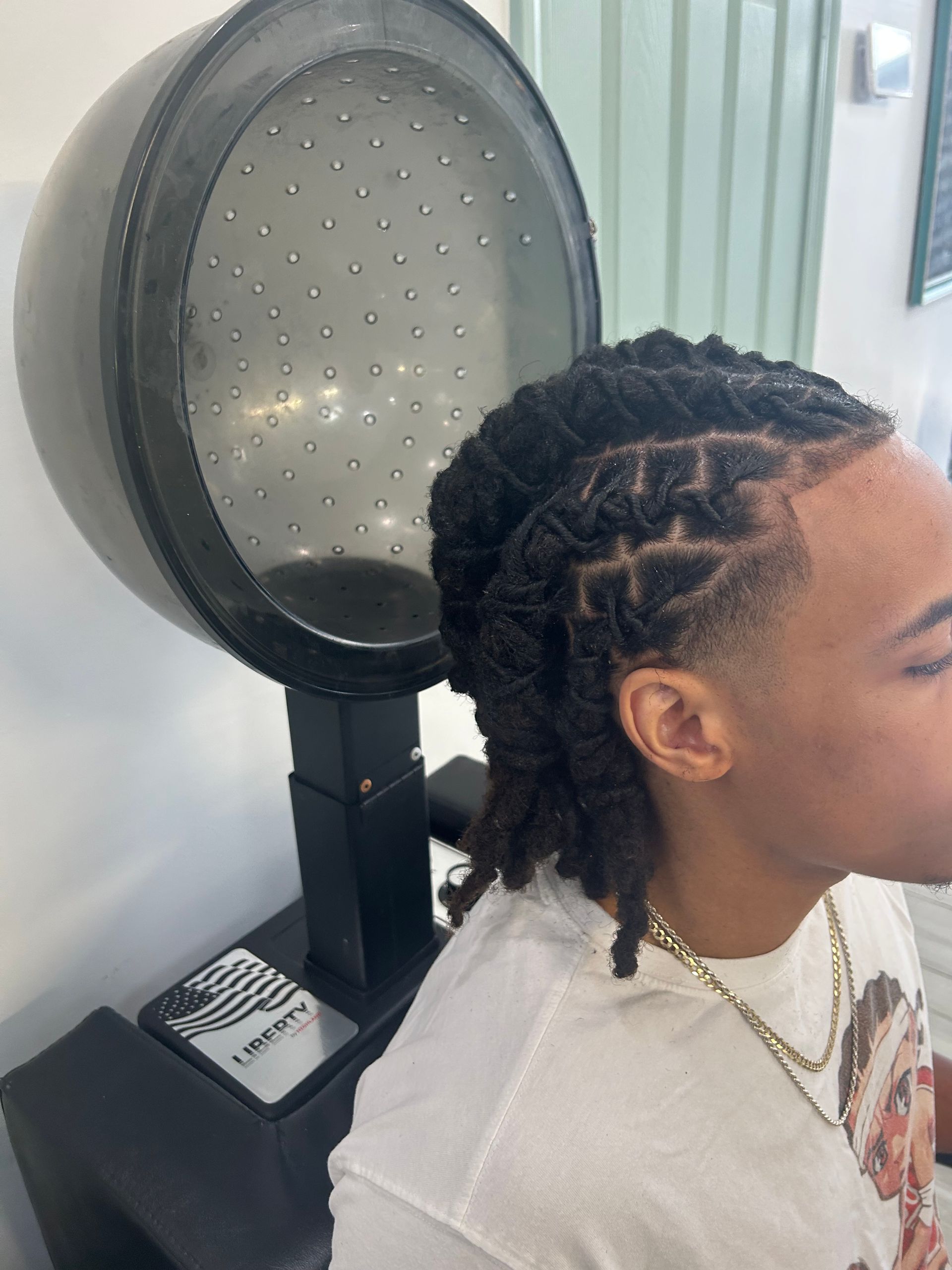 Person seated under a hooded salon dryer, with braided hair and a chain necklace, in a bright salon.