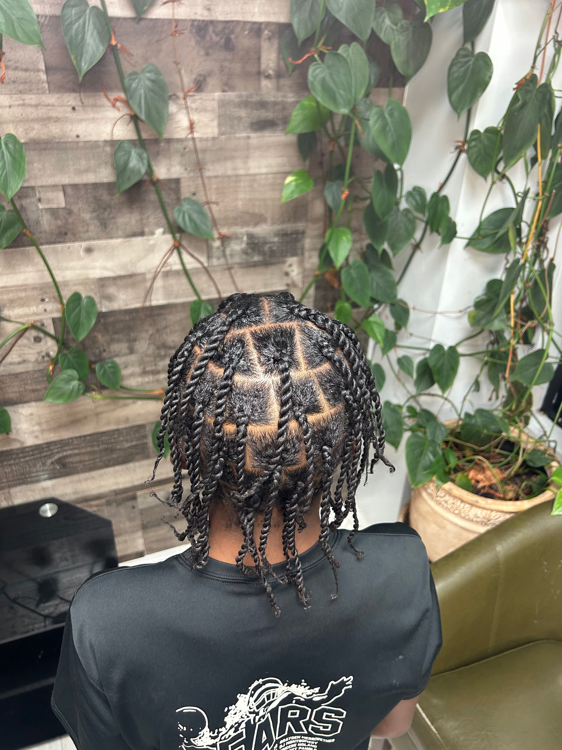 Person with braided hair seated outdoors against a leafy wall and wooden paneling.