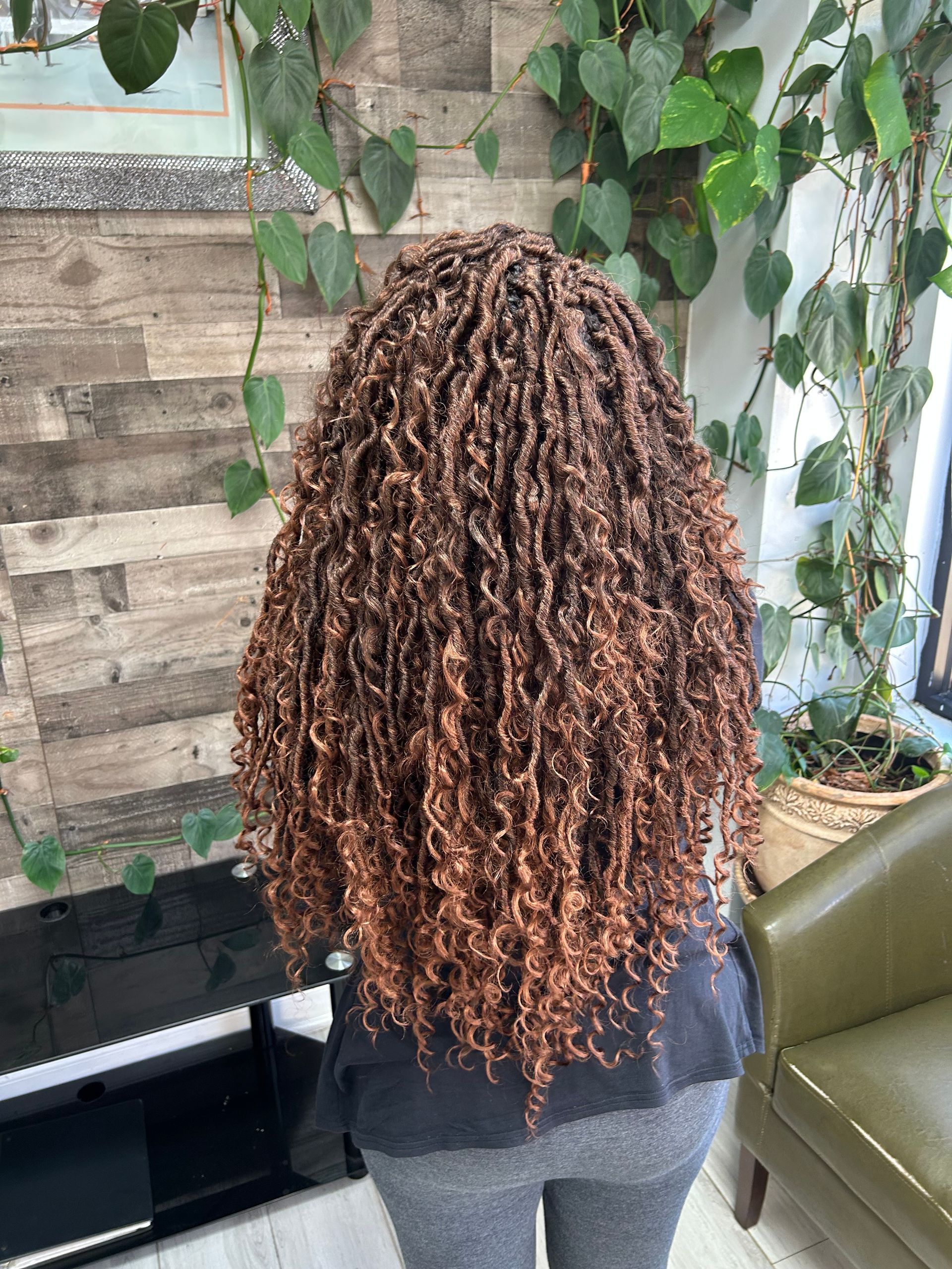Person with long curly brown hair standing indoors near a TV and green couch with hanging plants.