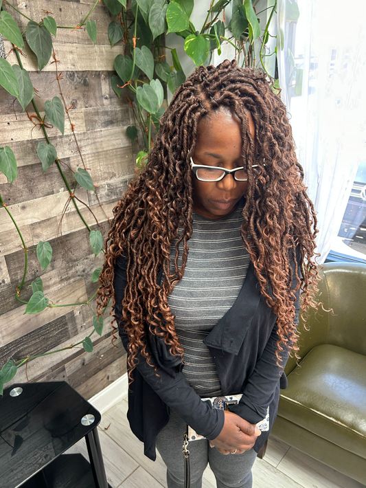Woman with long braids and glasses standing by a leafy brick wall, looking down with hands clasped.
