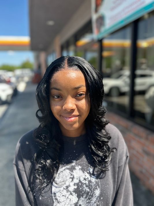 Smiling person standing outdoors in sunlight, with storefronts and parked cars behind them.