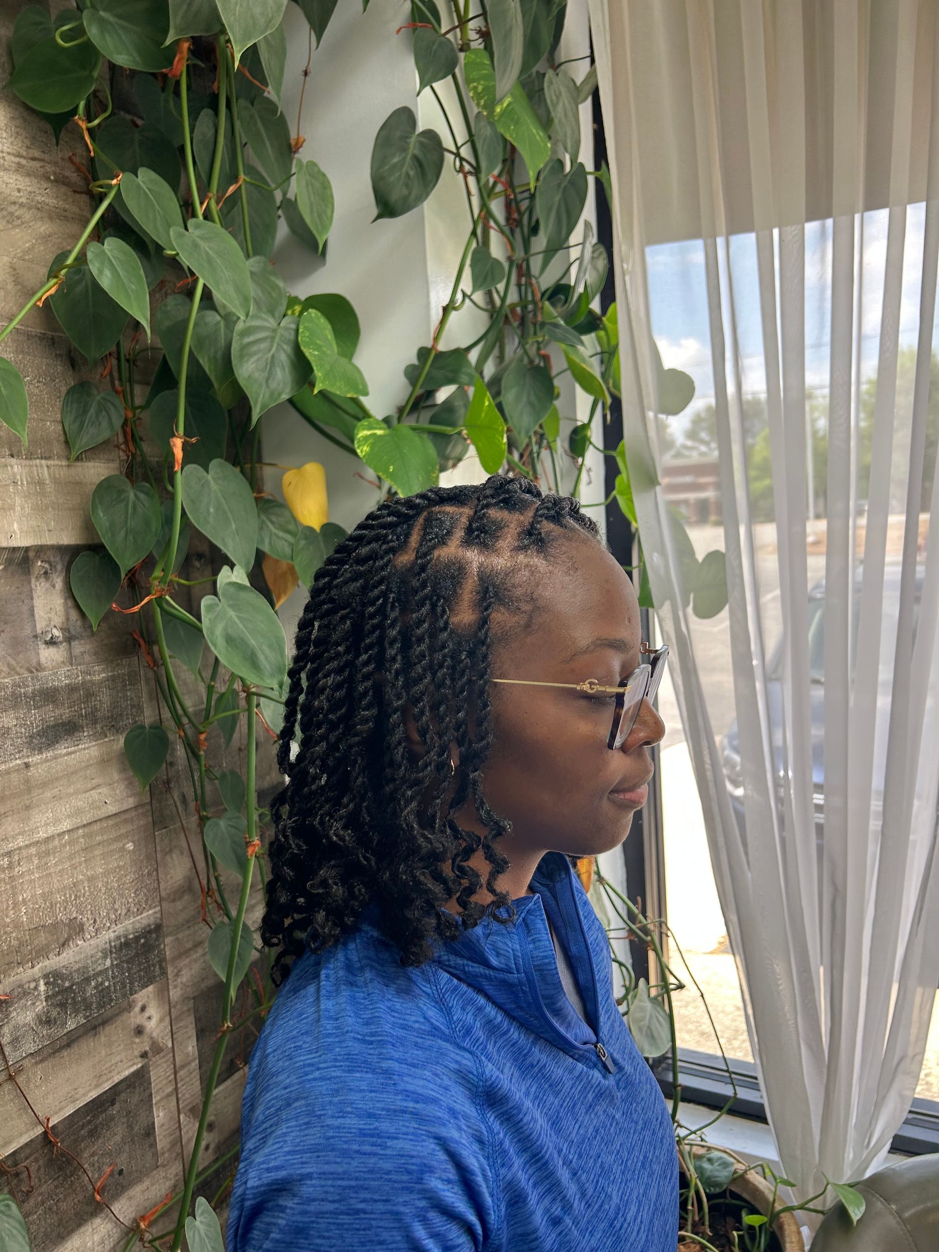 Person in blue jacket with braided hair, standing by ivy-covered wall and sheer curtains, looking out a window.