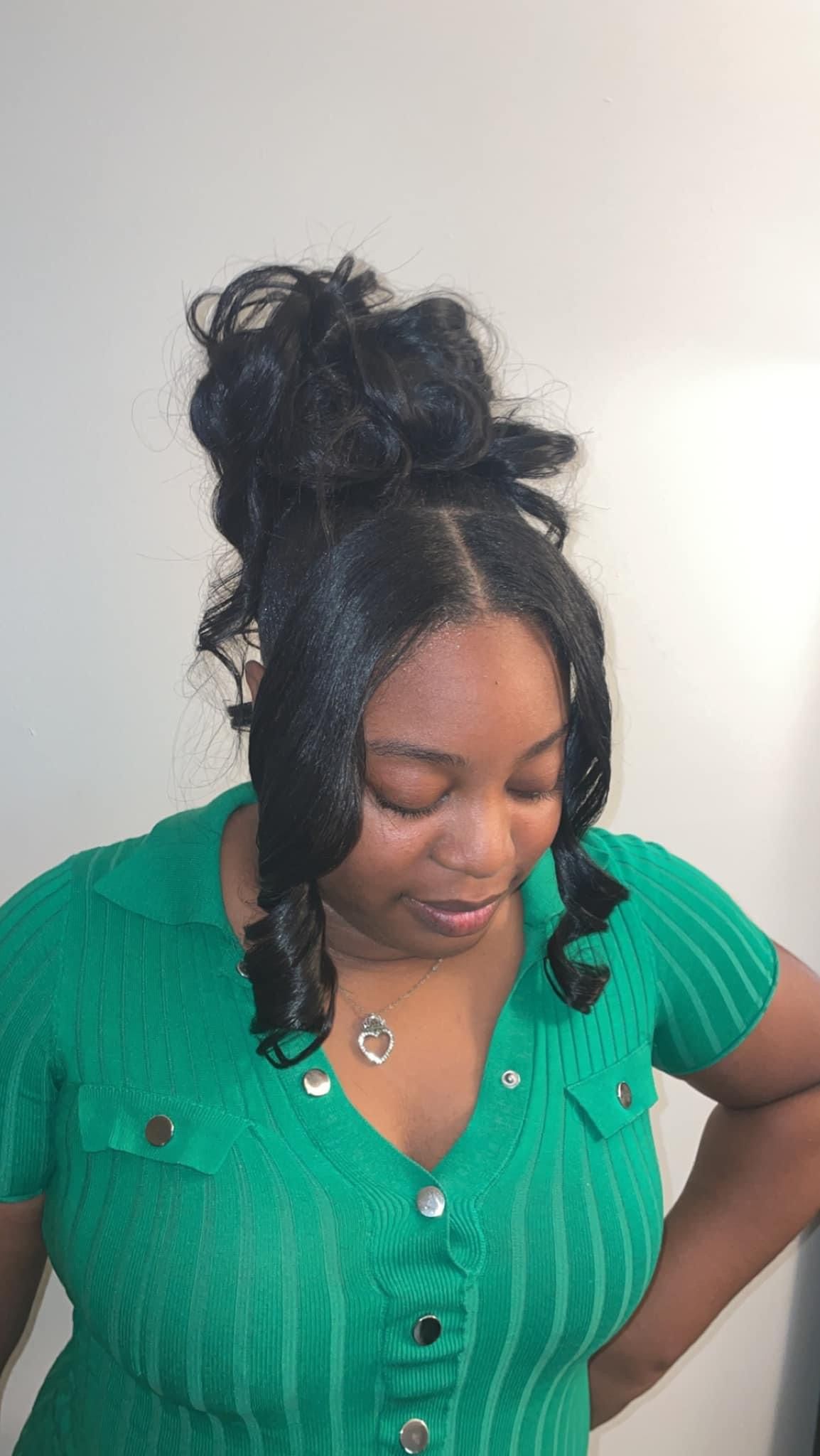Woman in a green top looking down with hair in a high bun, against a plain light wall.
