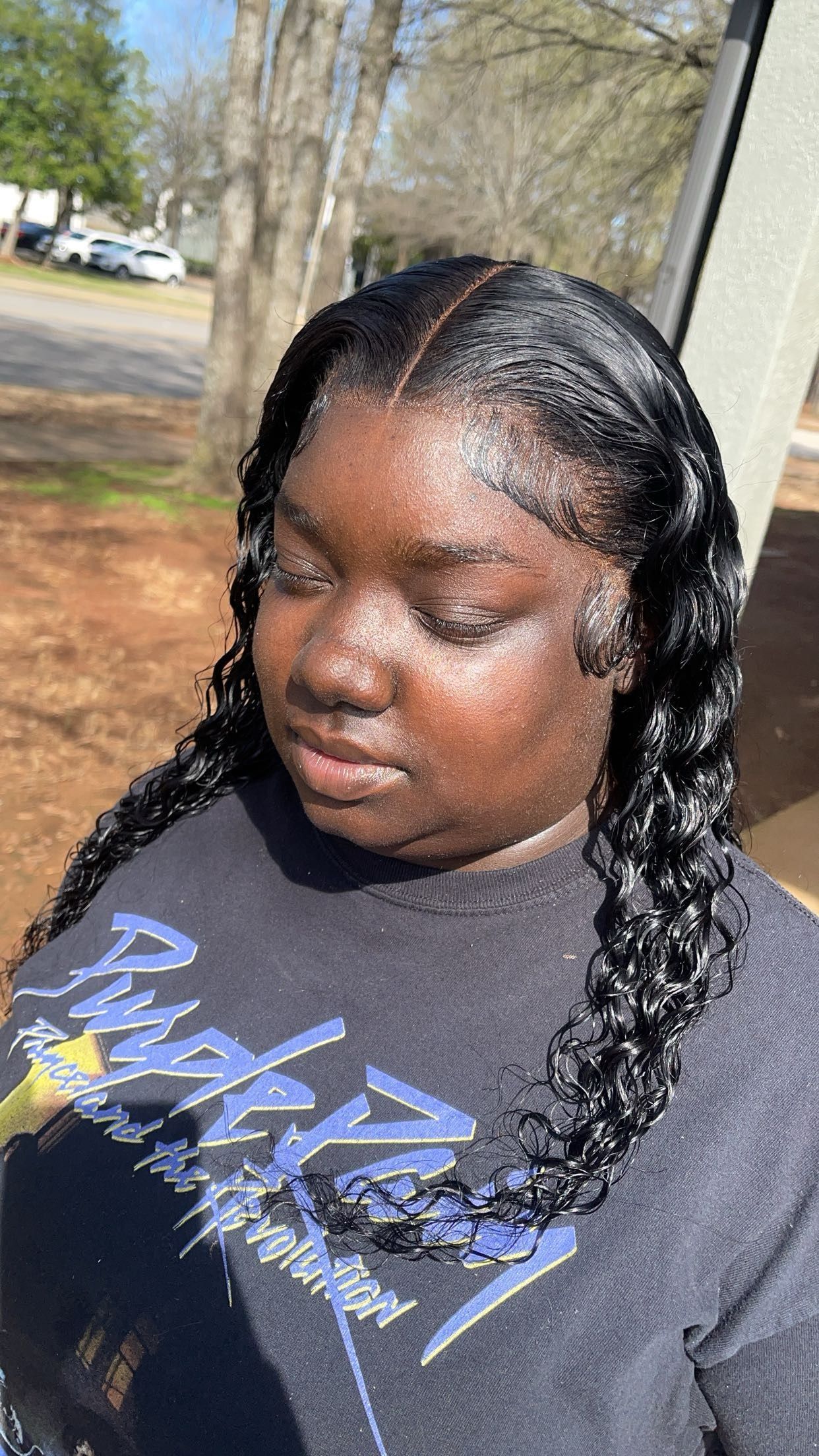 Person with braided hair wearing a black graphic T-shirt, standing outdoors in sunlight.
