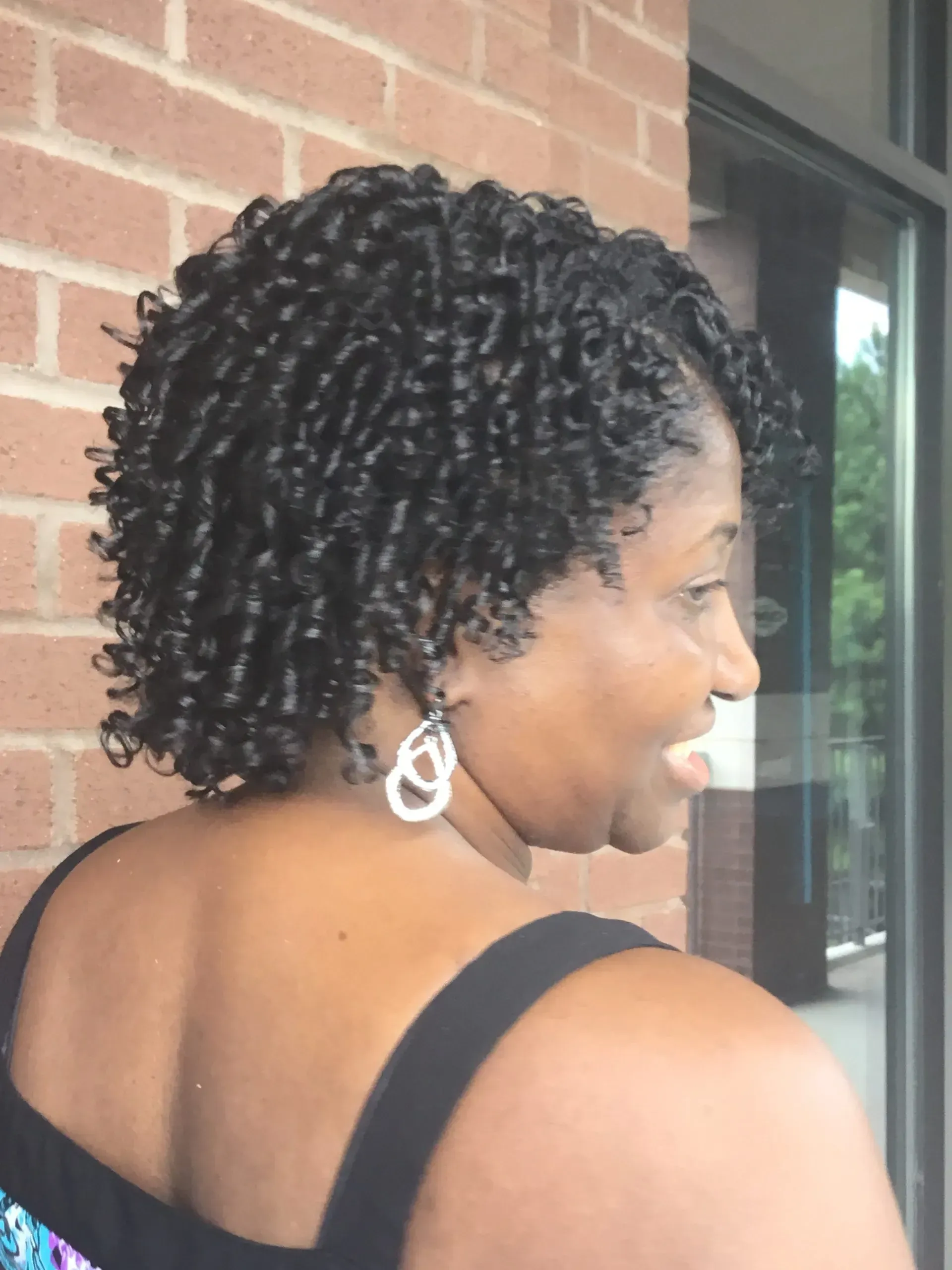 Woman with short curly hair and silver earrings, viewed from the side near a brick wall and window.