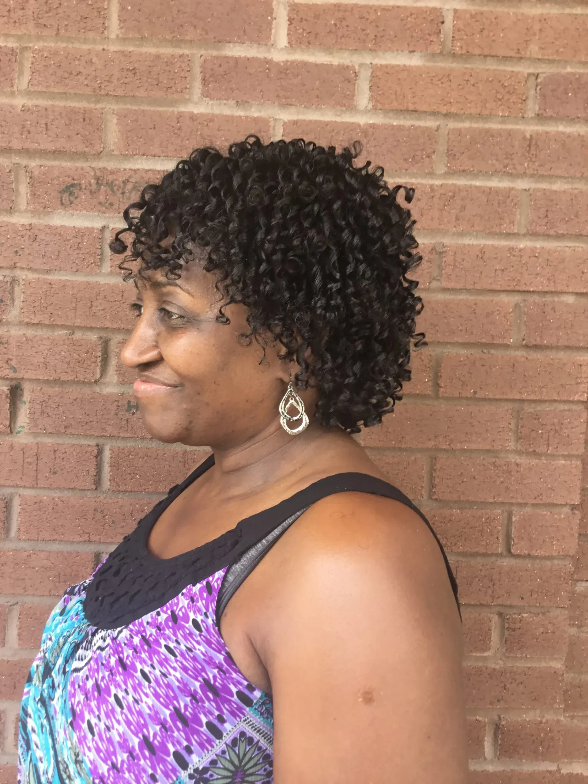 Profile view of a smiling woman in a purple patterned top against a brick wall