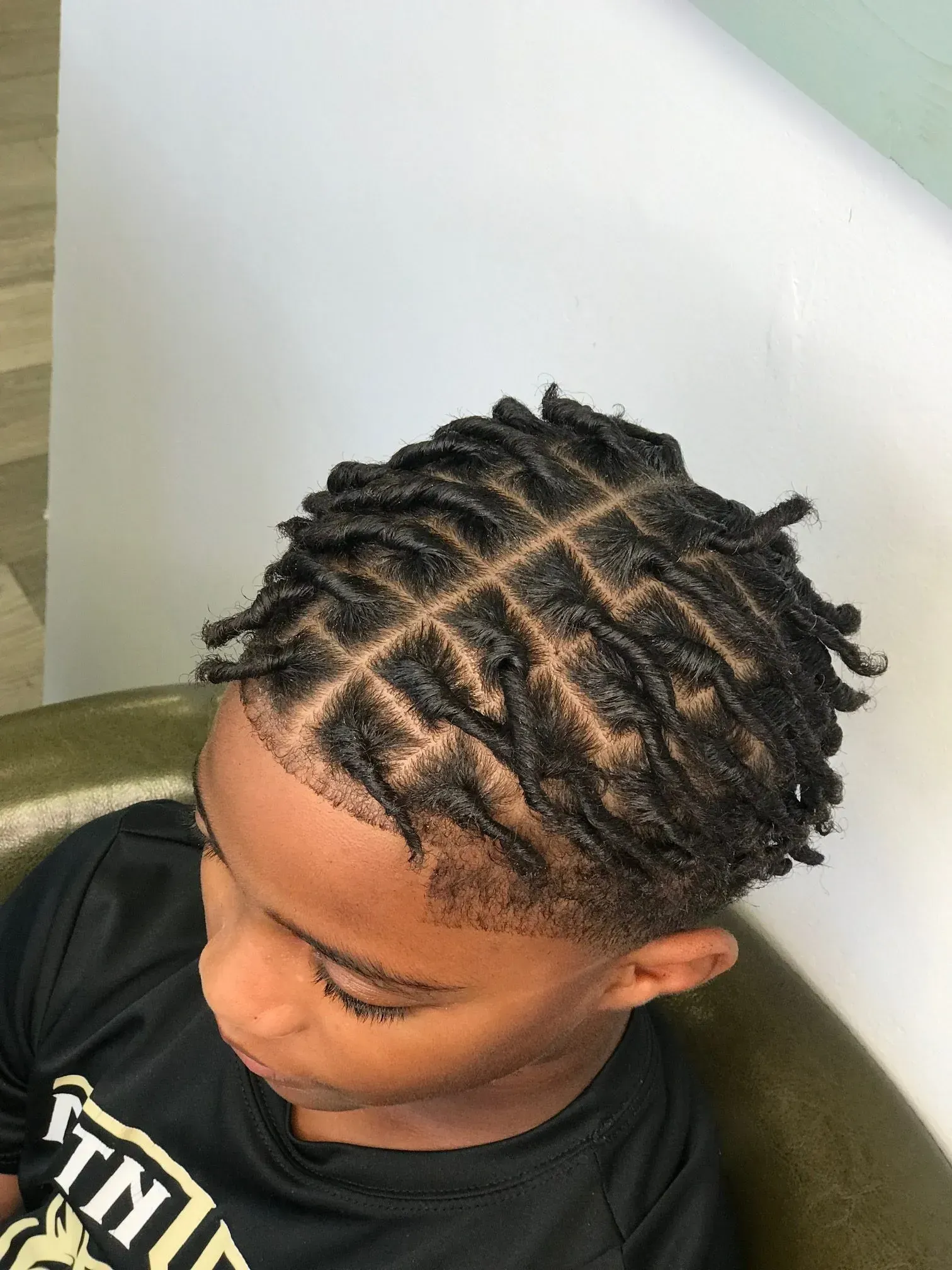 Top-down view of a child with neatly parted braided hair sitting on a chair by a white wall.