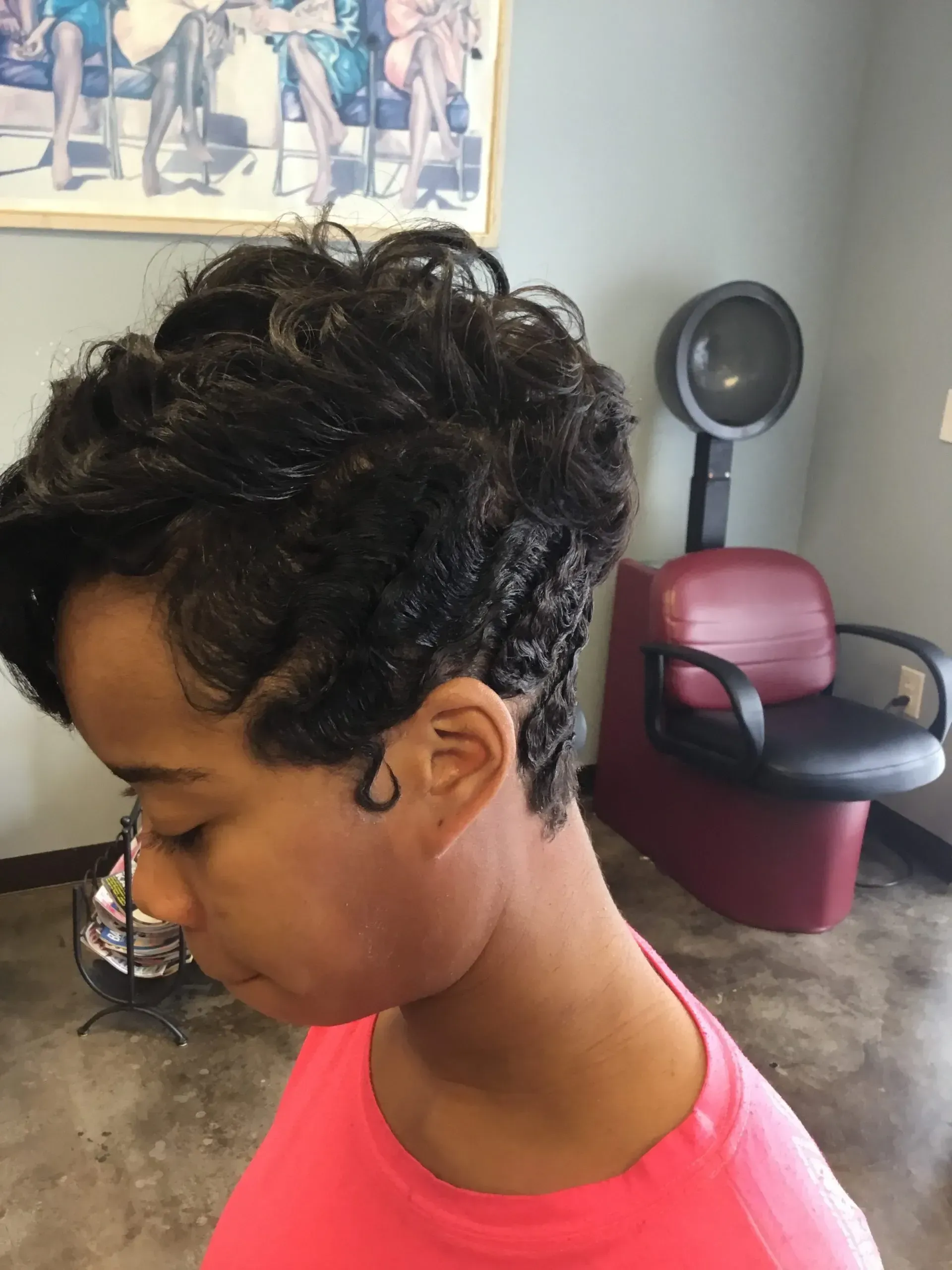 Side profile of a person with short curly hair in a pink shirt, indoors near a salon chair and wall poster.