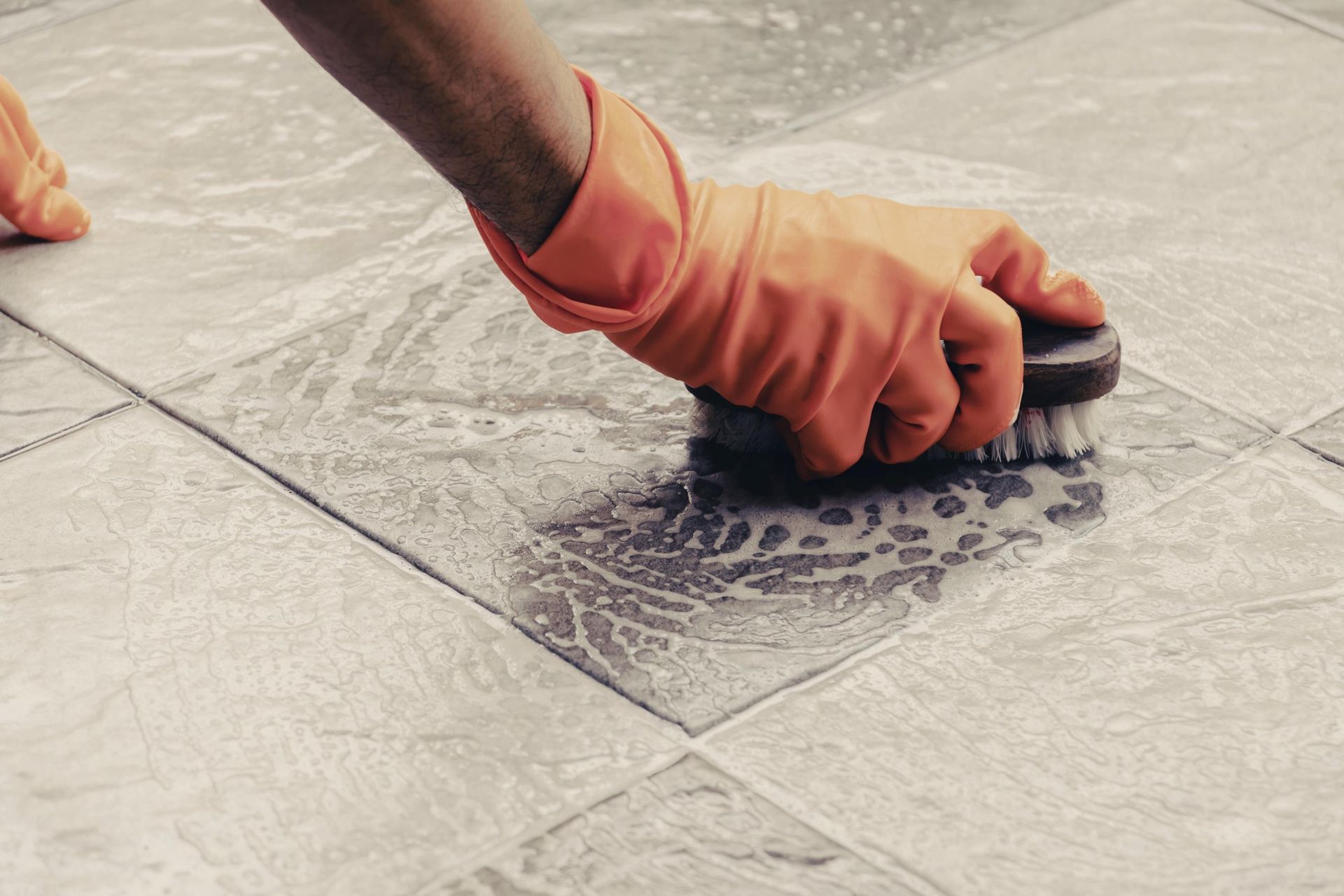 A person wearing orange gloves is cleaning a tile floor with a brush.