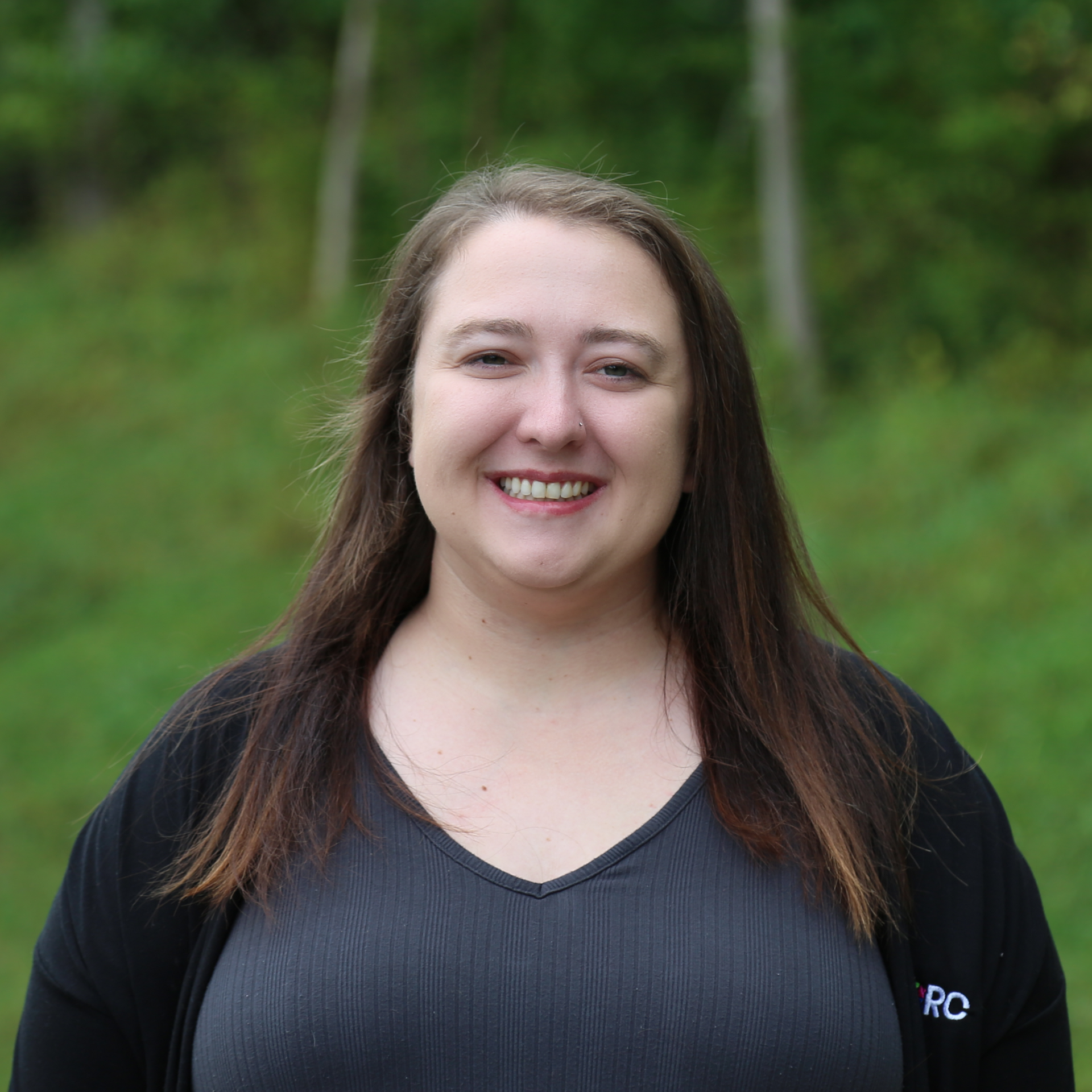 Woman with long brown hair smiles at the camera, wearing a black top. Outdoors with a green background.