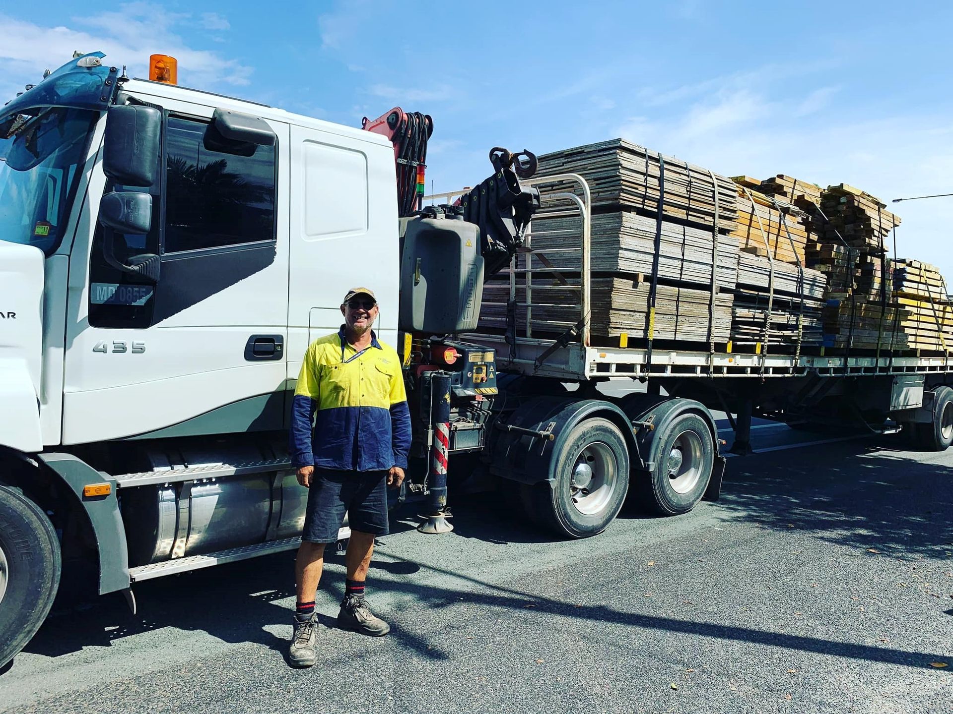 Worker standing beside white flatbed truck loaded with pallets on a gravel lot — Trucking Crane Trucks in Warana, QLD