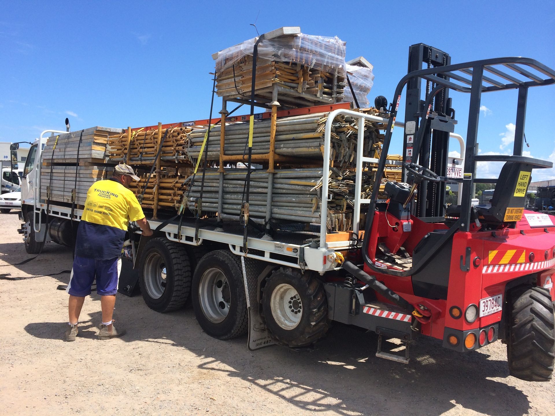 Forklift loading stacked pallets onto a flatbed truck at a sunny industrial site — Trucking Crane Trucks in Warana, QLD