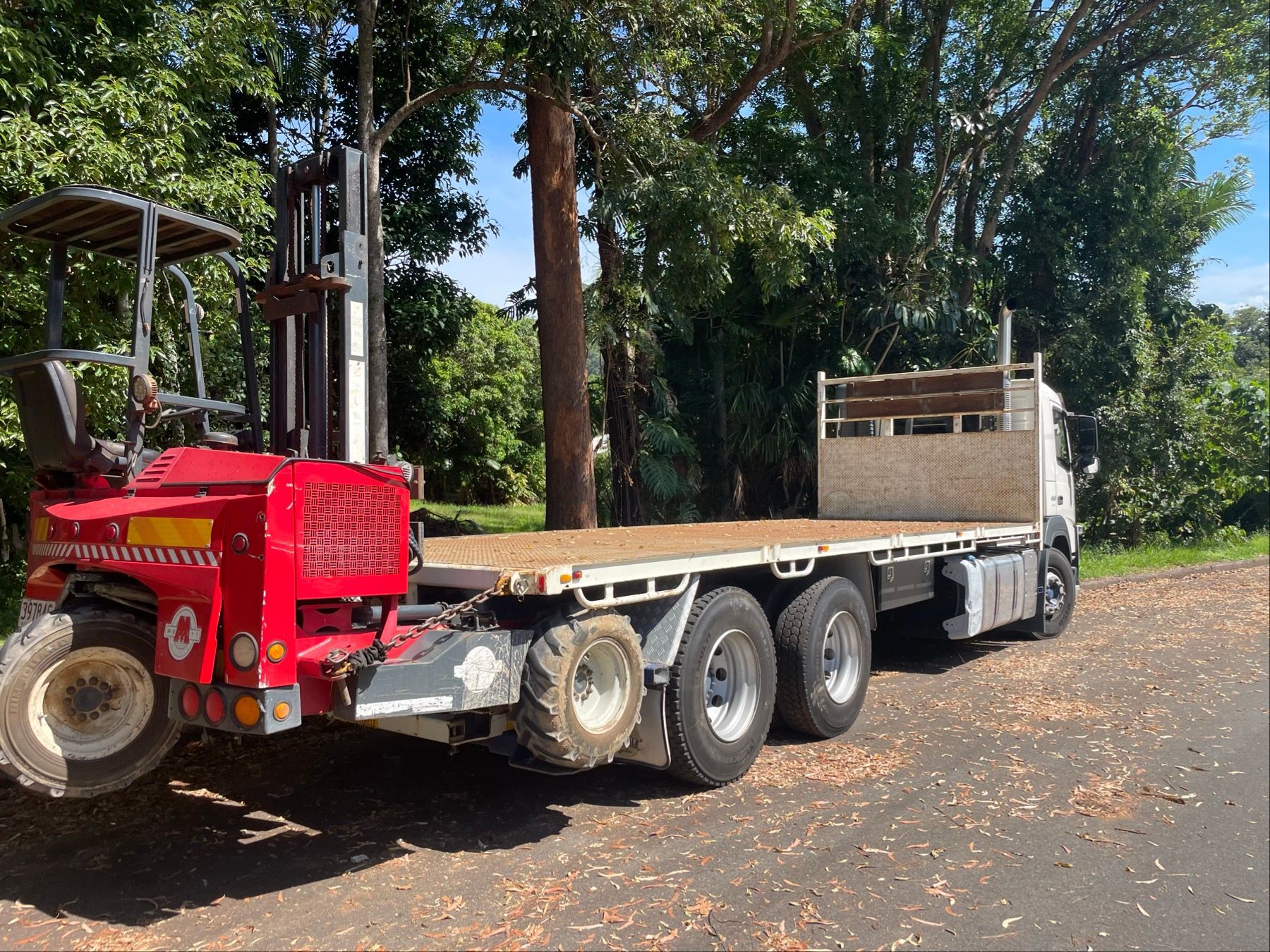 Red flatbed truck trailer parked on a dirt road under trees, carrying a forklift. — Trucking Crane Trucks in Caboolture, QLD